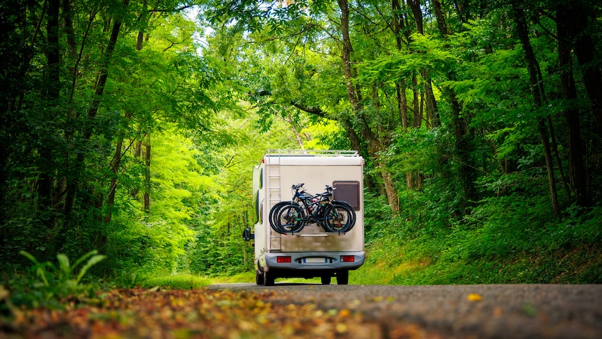 RV driving through a lush green forest with bicycles mounted on the back, symbolizing outdoor adventure and road travel.
