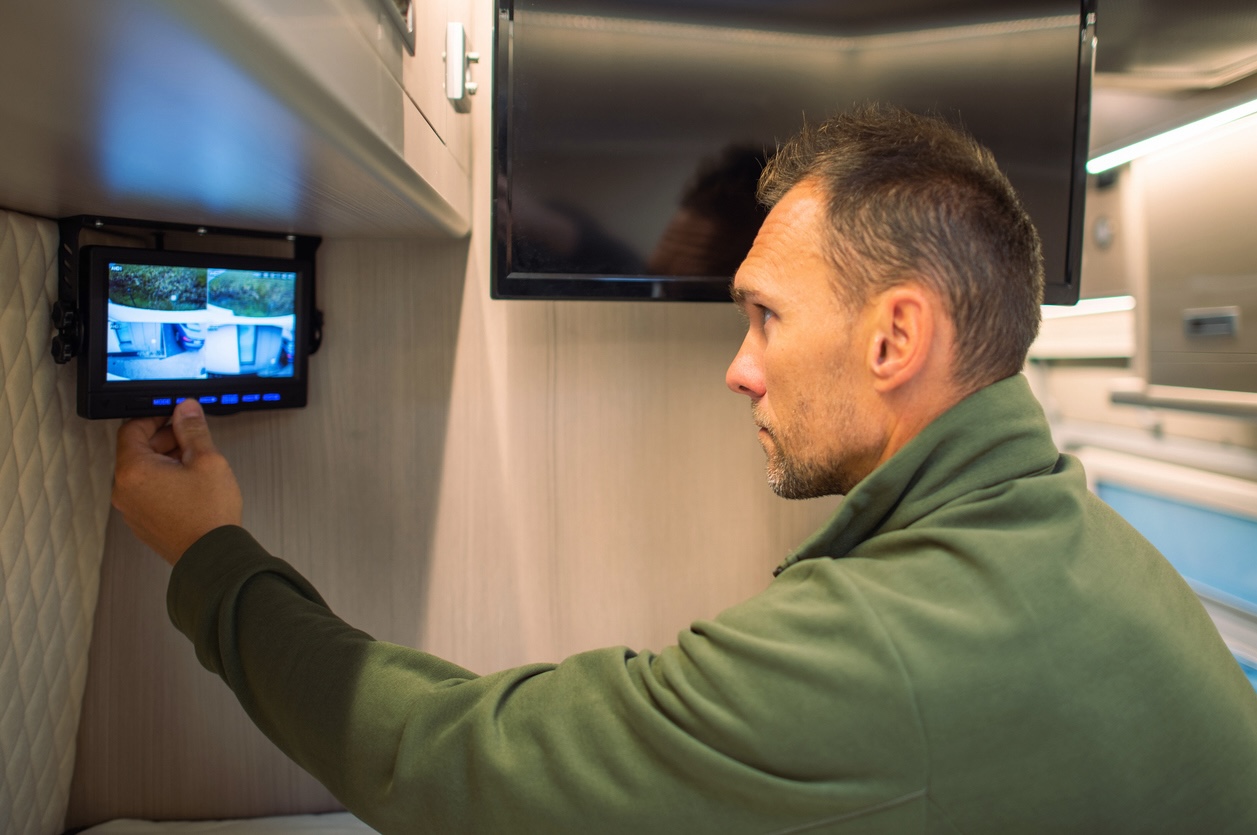 Man adjusting a security monitor inside a modern RV camper, showing outdoor camera views.