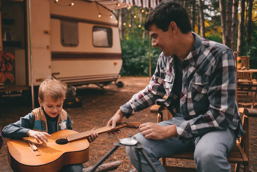 Father and son playing guitar together outside an RV at a peaceful wooded campsite.