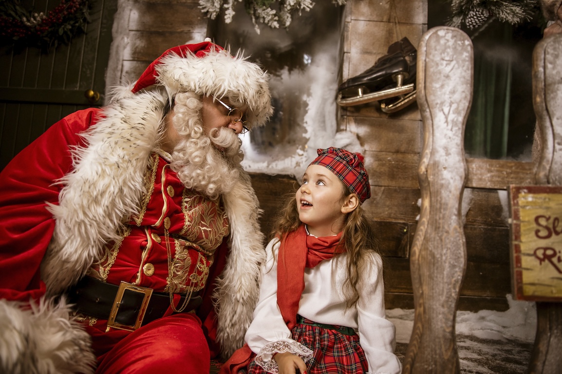 Santa Claus and a young girl sharing a festive moment in a rustic barn decorated for Christmas.