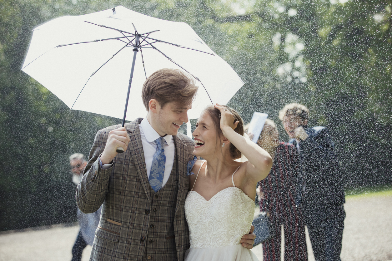 Bride and groom smiling beneath an umbrella during a rainy outdoor wedding